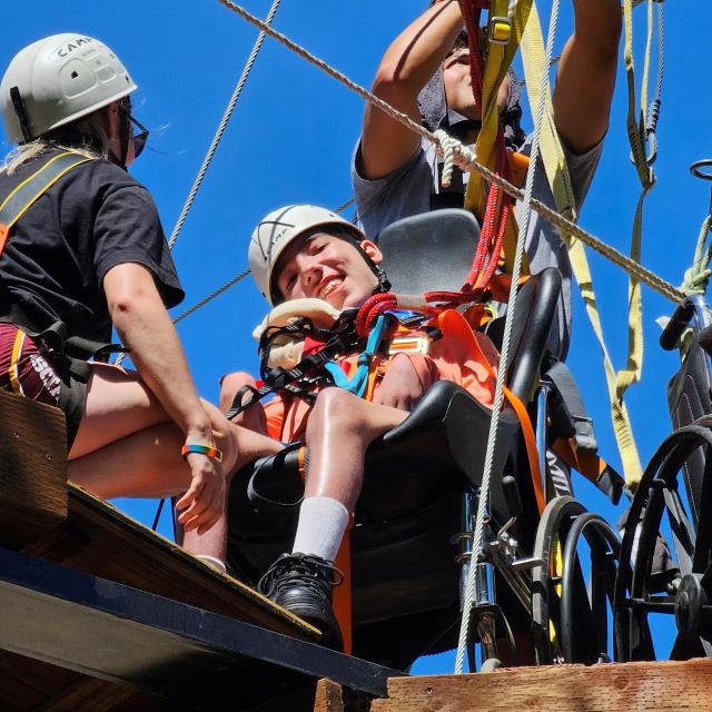 Teen boy in wheelchair featured on platform of high ropes course preparing to zipline out of his chair and fly through the air. Two staff members check the ropes and prepare him for the adventure. 